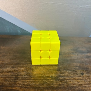 May include: A bright yellow Rubik's Cube puzzle, a classic brain teaser. The cube is composed of smaller squares in a grid pattern, each with a solid yellow color. The cube is sitting on a dark brown wooden surface.