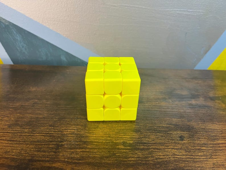 May include: A bright yellow Rubik's Cube puzzle, a classic brain teaser. The cube is composed of smaller squares in a grid pattern, each with a solid yellow color. The cube is sitting on a dark brown wooden surface.