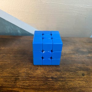 May include: A bright blue Rubik's Cube puzzle, a classic brain teaser. The cube is composed of nine squares on each of its six faces, each square a solid shade of blue. The cube sits on a dark wooden surface.