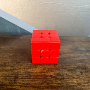 May include: A bright red Rubik's Cube, a classic puzzle toy, sits on a wooden surface. The cube is composed of smaller squares, each a vibrant red. The background features a wall with geometric shapes.