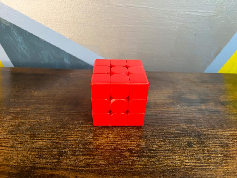 May include: A bright red Rubik's Cube, a classic puzzle toy, sits on a wooden surface. The cube is composed of smaller squares, each a vibrant red. The background features a wall with geometric shapes.