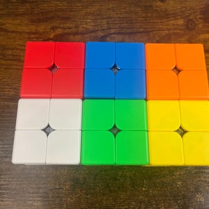 Peut inclure: Un jouet de puzzle coloré avec six ensembles de carrés de deux par deux. Les couleurs sont rouge, bleu, orange, blanc, vert et jaune. Les carrés sont disposés en forme rectangulaire sur une surface en bois.