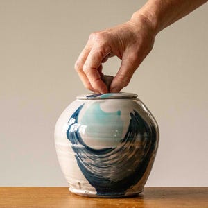 Può includere: Un vaso di ceramica con coperchio, con una base bianca con pennellate blu e turchese. Il vaso è rotondo con una forma leggermente bombata. Una mano si allunga per sollevare il coperchio. Il vaso è su una superficie di legno.