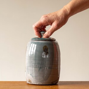 Op de afbeelding: Een handgemaakte keramische pot met een deksel, in tinten blauw en wit. De pot heeft een afgeronde vorm en een glanzende afwerking. Een hand reikt om het deksel op te tillen. De pot staat op een houten oppervlak.