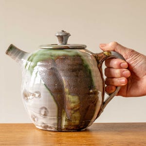 Op de afbeelding: Een keramische theepot met een deksel, tuit en handvat. De theepot heeft een afgeronde vorm en is geglazuurd met een mix van kleuren, waaronder wit, bruin en groen. De theepot staat op een houten oppervlak.