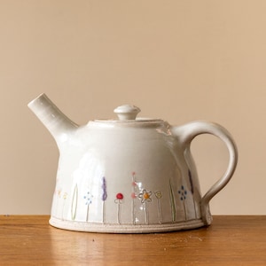 May include: A white ceramic teapot with a floral design. The teapot has a round body and a spout on the side. The handle is on the right side of the teapot.