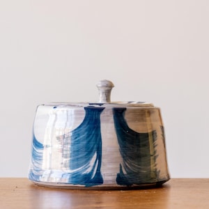 May include: A ceramic lidded jar with a rounded rectangular shape. The jar is painted with a white and blue abstract design. The lid has a small knob. The jar sits on a wooden surface.