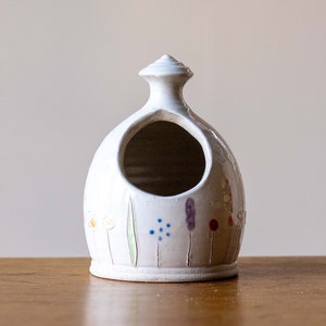 Può includere: Saliera in ceramica bianca con una piccola apertura nella parte superiore e un design floreale in tonalità di verde, blu, viola, rosso e giallo.