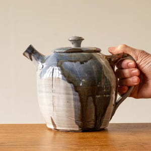 Könnte beinhalten: Eine handgefertigte Keramik-Teekanne mit Deckel, mit einer einzigartigen Glasur in Weiß-, Blau- und Brauntönen. Die Teekanne hat einen gebogenen Ausguss und einen Griff, perfekt zum Servieren von Tee. Die Teekanne steht auf einer Holzoberfläche.