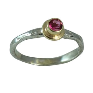 Könnte beinhalten: Ein Silberring mit einer goldenen Lünette, die einen runden, rosa Edelstein hält. Der Ring hat ein gehämmertes Band und eine polierte Oberfläche. Der Edelstein hat eine leuchtend rosa Farbe.