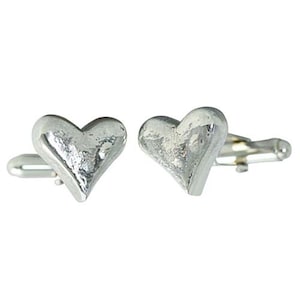 May include: A pair of silver-colored heart-shaped cufflinks. The cufflinks have a textured surface and a classic T-bar closure. The heart design is slightly rounded, giving them a three-dimensional appearance.