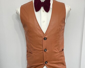 May include: A tan leather vest with three dark buttons and two pockets. A burgundy velvet bow tie is paired with a white dress shirt. The outfit is displayed on a mannequin.