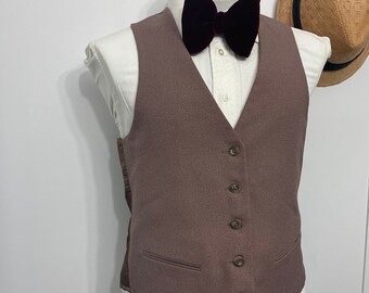 May include: A brown vest with a five-button closure and two front pockets. A dark burgundy velvet bow tie is paired with a white collared shirt. A straw hat with a black band hangs in the background.