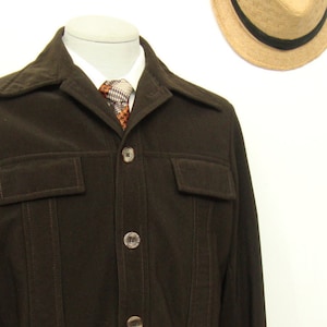 Może przedstawiać: Ciemnobrązowa sztruksowa kurtka z zapięciem na guziki, dwiema kieszeniami na piersi i kołnierzem z klapami. Kurtka jest prezentowana na manekinie z wzorzystym krawatem i białą koszulą. Słomkowy kapelusz z czarną opaską jest widoczny w tle.