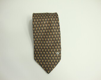 May include: A patterned necktie with a repeating geometric design in shades of brown, black, and tan. The tie has a pointed end and a small silver emblem near the bottom. The tie is made of a woven fabric.