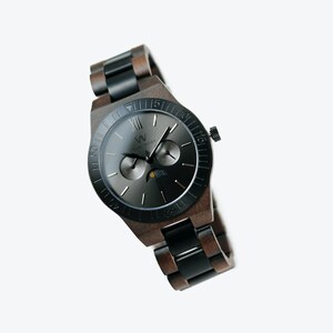 May include: A black and brown wooden wristwatch with a black face and silver hands. The watch has a moon phase complication and a date window. The watch is made by the brand "Wood & Watch".