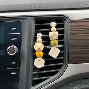 Pode incluir: Dois ambientadores de carro pendurados numa grelha de ventilação. Cada ambientador apresenta uma combinação de contas de madeira, bolas de feltro e formas geométricas em tons naturais, amarelos e verdes. Os ambientadores são fixados com uma presilha prateada.