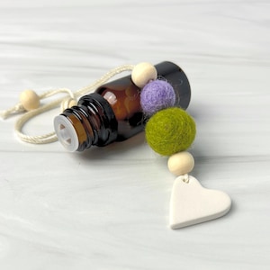 Puede incluir: Una botella de vidrio ámbar para aceites esenciales con una tapa de plástico blanca, una cuerda color crema y un adorno decorativo de fieltro y cuentas de madera. El adorno incluye una bola de fieltro verde, morada y un colgante de cerámica blanco en forma de corazón.