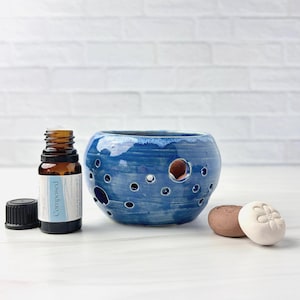Op de afbeelding: Een blauwe keramische etherische olie diffuser met meerdere gaten, een bruine glazen fles met het label "Composed" en twee kleine, ronde stenen. De diffuser heeft een glanzende afwerking en staat op een wit oppervlak.