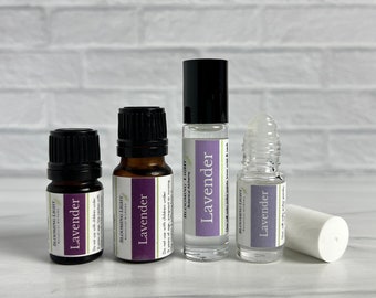 Op de afbeelding: Een assortiment lavendel etherische olie producten. De set bevat kleine glazen flesjes met zwarte doppen en etiketten, een rollerflesje en een wit cilindrisch object. De etiketten lezen "Lavender" en "Blooming Light Botanical Alchemy".