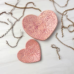 May include: Two heart-shaped trinket dishes in a pinkish-bronze hue, adorned with a rose pattern. The larger dish is approximately 6 inches across, while the smaller one is about 3 inches. Decorative brown paper shreds are scattered around the dishes.