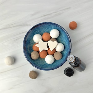 Puede incluir: Vista superior de un cuenco azul lleno de bolas de fieltro blancas, marrones y naranjas, junto con piezas de fieltro en forma de estrella y corazón. Una pequeña botella de aceite esencial marrón y una tapa negra están al lado del cuenco.