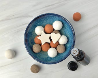 May include: A top-down view of a blue bowl filled with white, brown, and orange felt balls, along with star and heart-shaped felt pieces. A small brown essential oil bottle and a black cap are next to the bowl. The objects are on a white marble surface.
