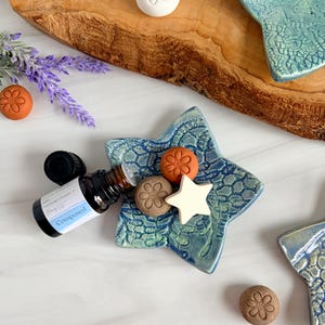 May include: A star-shaped blue ceramic dish with a textured lace pattern, holding a small amber glass essential oil bottle labeled Composed, a white star-shaped wax melt, and two small round terracotta-colored floral wax discs.