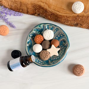May include: A teal ceramic dish holds several small clay shapes, including circles with flower imprints and a star. Beside the dish lies a brown glass bottle with a black cap and a label reading Composed, along with loose clay pieces.