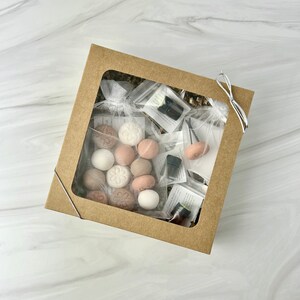 May include: A brown cardboard box with a clear window showing a variety of items, including small round scented discs in various shades of brown and white, small glass bottles with dark liquid, and a small white mesh bag with a metal hook. The box is tied with a white ribbon.