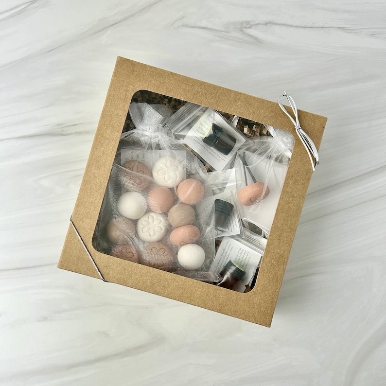 May include: A brown cardboard box with a clear window showing a variety of items, including small round scented discs in various shades of brown and white, small glass bottles with dark liquid, and a small white mesh bag with a metal hook. The box is tied with a white ribbon.