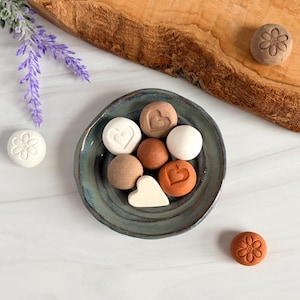 May include: A small, round, blue-green ceramic dish holds several clay-like spheres and shapes in shades of white, tan, brown, and terracotta. Some pieces feature embossed heart or flower designs. Additional loose pieces and lavender sprigs are nearby.
