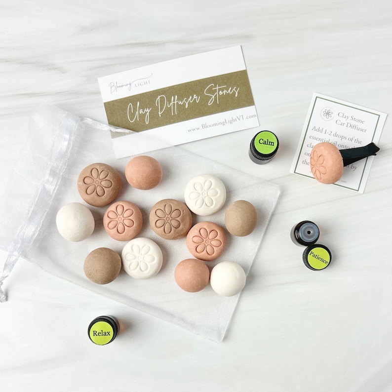 May include: A set of 12 clay diffuser stones in beige, brown, and white with a flower design. The stones are in a white mesh bag. Three small bottles of essential oil are labeled "Relax", "Calm", and "Patience".