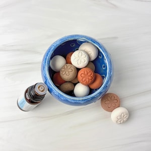Puede incluir: Un cuenco de cerámica azul lleno de piedras de varios colores con flores en relieve. Junto al cuenco hay una pequeña botella de aceite esencial marrón con una etiqueta blanca. Las piedras son de color blanco, marrón y naranja.