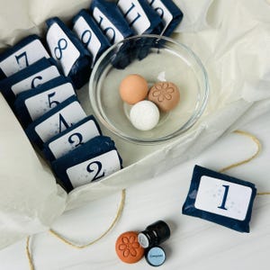 May include: A set of numbered dark blue wrapped packages from 1 to 12, a clear glass bowl containing three small round spheres, and a small dark brown bottle with a blue label. One sphere is white, one is tan, and one is a light brown with a flower design.