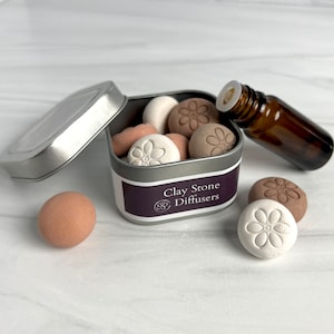 May include: A silver tin with a lid open, revealing a set of small clay stone diffusers in various shades of brown and white. The diffusers are shaped like flowers with a raised design. The tin has a label that reads "Clay Stone Diffusers". A small brown glass bottle with a dropper is also visible.