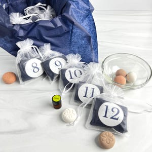 Könnte beinhalten: Ein Adventskalender mit nummerierten Beuteln, die mit dunkelblauen Artikeln gefüllt sind. Enthält eine Glasschale mit Eiern, eine kleine Flasche mit der Aufschrift "Calm" und dekorative Badebomben. Die Beutel befinden sich in einer blau ausgekleideten Box.