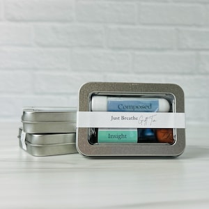 May include: A rectangular metal gift tin containing a lip balm and two small tubes. The lip balm is labeled "Composed", and the tubes are labeled "Insight". The tin is wrapped with a white band that reads "Just Breathe Gift Tin".