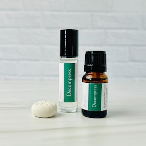 Puede incluir: Se muestran dos botellas de vidrio con tapas negras y etiquetas verdes que dicen "Decompress". Una es una botella roll-on, la otra una pequeña botella ámbar. También hay una pequeña piedra redonda de color blanquecino.