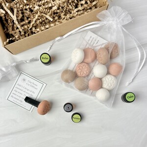 May include: A set of 12 clay stone car diffusers in a white organza bag. The diffusers are in various shades of brown, beige, and white. The bag is sitting on a white surface. There are four small bottles of essential oils labeled "Relax", "Calm", "Patience", and a fourth bottle with an unreadable label. The image also includes a clay stone diffuser attached to a metal clip.