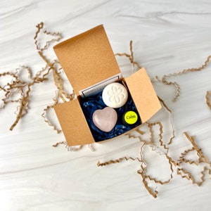 Könnte beinhalten: Eine kleine, offene Pappschachtel mit einem herzförmigen Rosenquarzstein, einer runden weißen Seife mit Blumendesign und einem kleinen gelben Behälter mit der Aufschrift "Calm". Die Schachtel ist mit blauem Seidenpapier ausgekleidet.