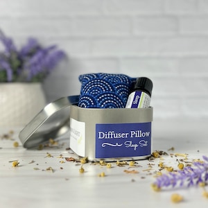 May include: A silver tin with a blue and white patterned diffuser pillow and a small bottle of essential oil labeled "Sleep". The tin has a label that reads "Diffuser Pillow Sleep Set".