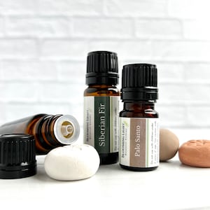 May include: Two amber glass bottles with black caps and labels that read "BLOOMING LIGHT Siberian Fir" and "BLOOMING LIGHT Palo Santo". The bottles are sitting on a white surface with two small ceramic stones.