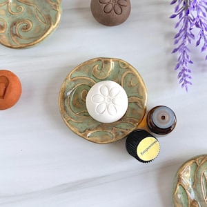 May include: A white circular diffuser stone with a flower relief pattern sits on a round, green and gold glazed ceramic dish. Nearby are a small brown glass essential oil bottle with a black cap labeled Empowered and sprigs of purple lavender.