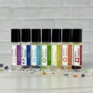 Puede incluir: Un conjunto de siete botellas de aceite esencial con rodillo, cada una etiquetada con el nombre y el símbolo de un chakra. Las botellas tienen tapas negras y cuerpos de vidrio transparente, llenas de aceites de colores y pequeños cristales.