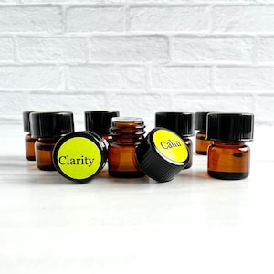 Op de afbeelding: Een groep van acht kleine amberkleurige glazen flesjes met zwarte dopjes en gele etiketten. De etiketten hebben de woorden "Calm" en "Clarity" erop gedrukt.