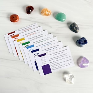 May include: A set of seven rectangular cards with chakra-themed text and symbols, arranged in a fan shape, accompanied by seven polished tumbled gemstones in red, orange, yellow, green, dark blue, indigo, and violet, plus one clear stone.