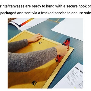 May include: A person using a red staple gun to attach the back of a framed print. The frame is light brown, and the back is a tan color. The text in the image says, "Framed prints/canvases are ready to hang with a secure hook on the back."