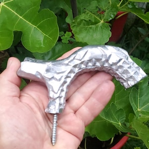 May include: A silver-colored, textured metal handle with a screw at the base. The handle has a unique, organic shape, possibly for a door or drawer. The background features green leaves.