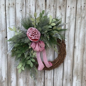 May include: A Christmas wreath with a pink flower and red and white striped ribbon. The wreath is made of natural materials and is decorated with greenery.